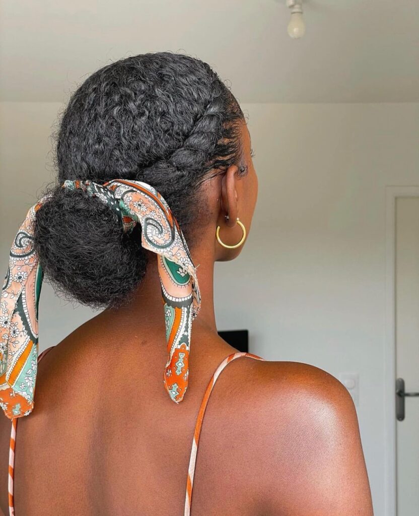 A woman viewed from the back with a side halo braid tucked into a low bun that is accessorized with a patterned orange and green silk scarf.