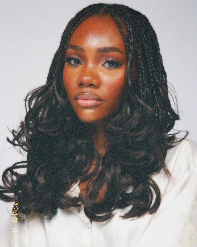 A woman with small black braids at the top of her head that flow into long, thick, and voluminous layered curls.