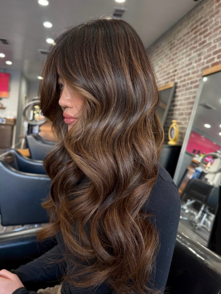 A woman in profile displays long, glossy dark hair styled in glossy waves with soft warm cinnamon highlights and curtain bangs.