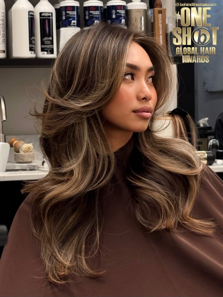 A young woman with long wavy dark brown hair featuring fine ash blonde highlights and soft face framing layers looks upward to the side.