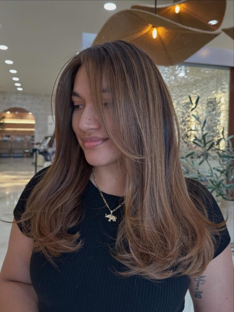 A woman with long light brown hair styled in soft outward curving layers and sweeping curtain bangs smiling gently in a salon.