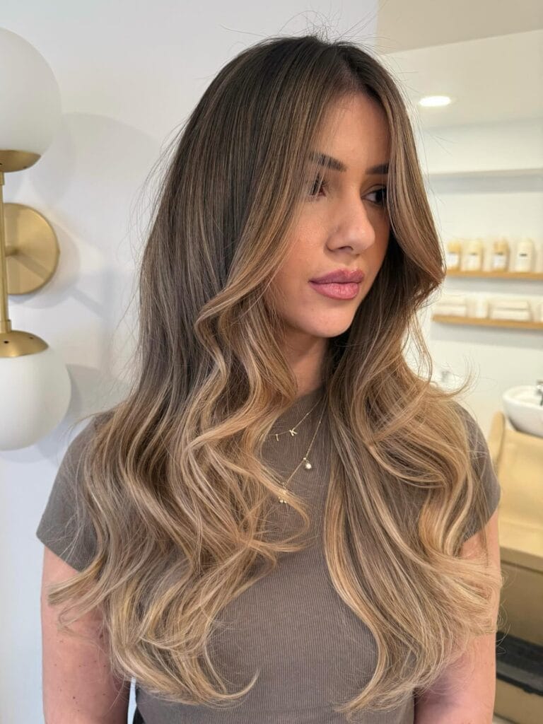 Long layered brunette hair with loose waves and golden beige face framing highlights.