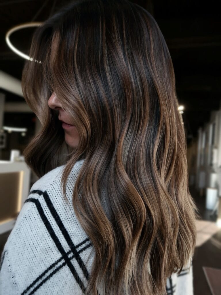 Long wavy brunette hair with soft caramel ribbon highlights and a glossy finish.