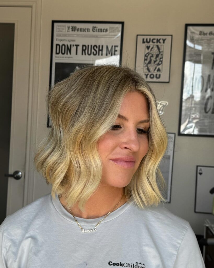 A woman with a shoulder-length blonde bob cut styled in loose, beachy waves looking down to the side.