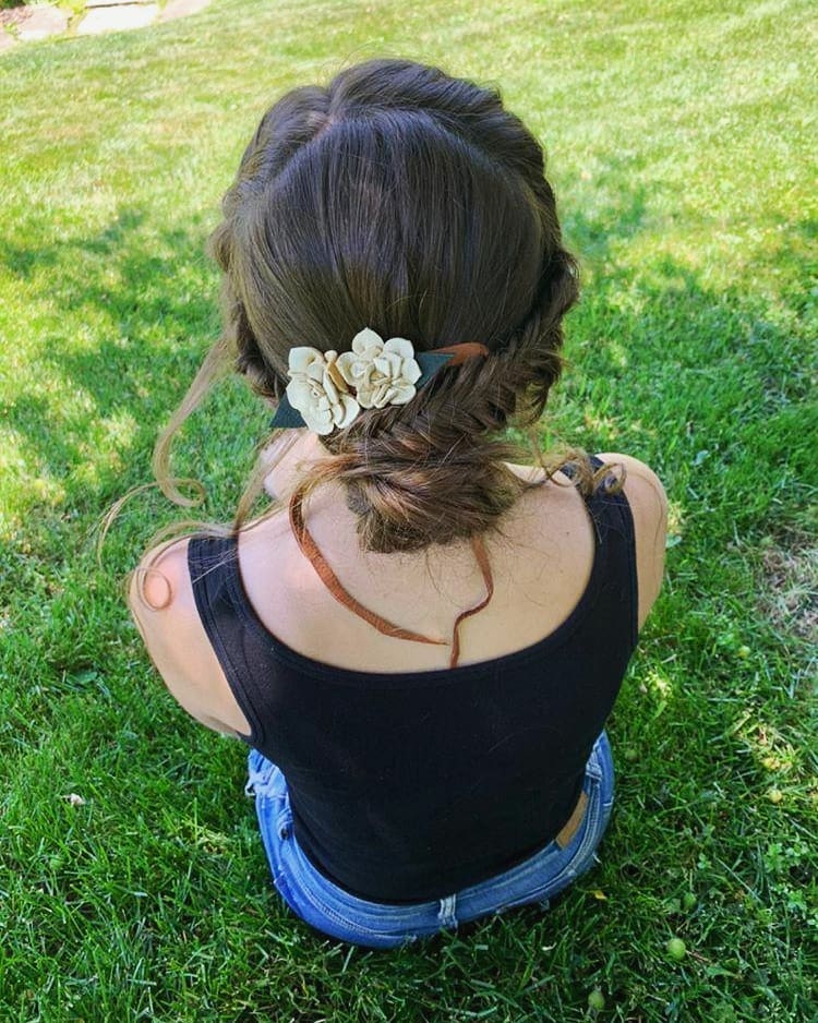 A rear view of a woman sitting on green grass with her brown hair styled in fishtail braids gathered into a low, messy bun decorated with cream flowers.
