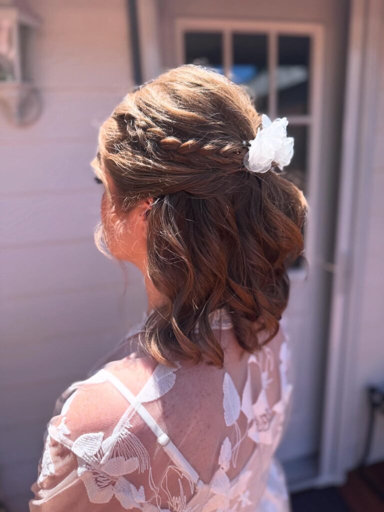 Short brunette hair styled in soft curls with braided half up sections and a white floral clip.
