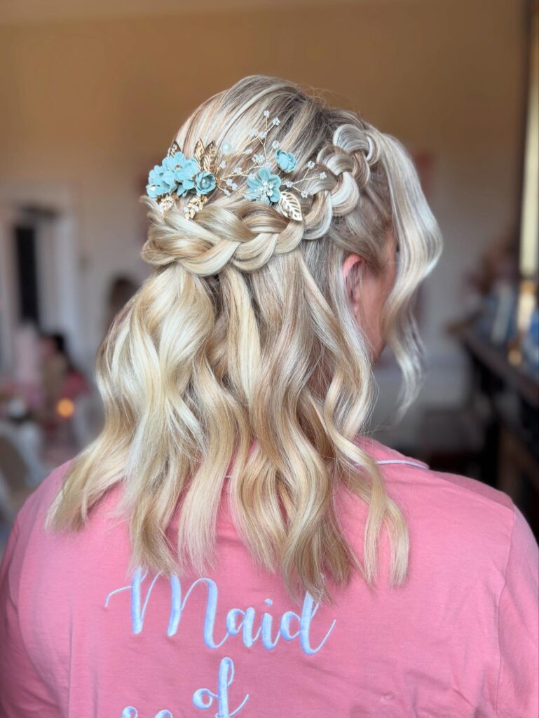 Blonde shoulder length hair styled in waves with crown braids and a blue floral accessory.