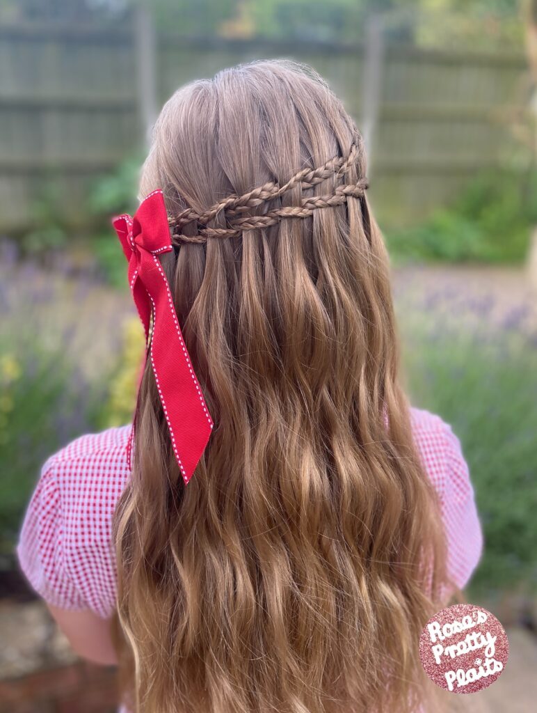 Long wavy light brown hair styled with crown braids and a red ribbon tied on the side.