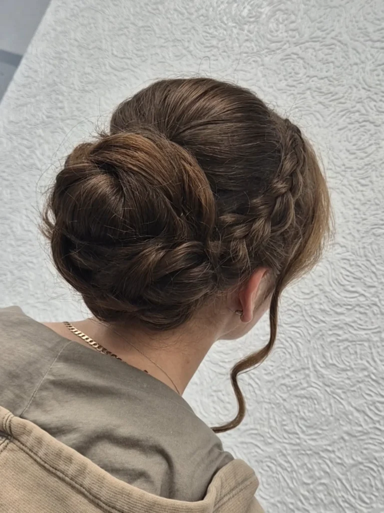 Smooth brunette braided low roll bun with a side braid and curled face framing strand.