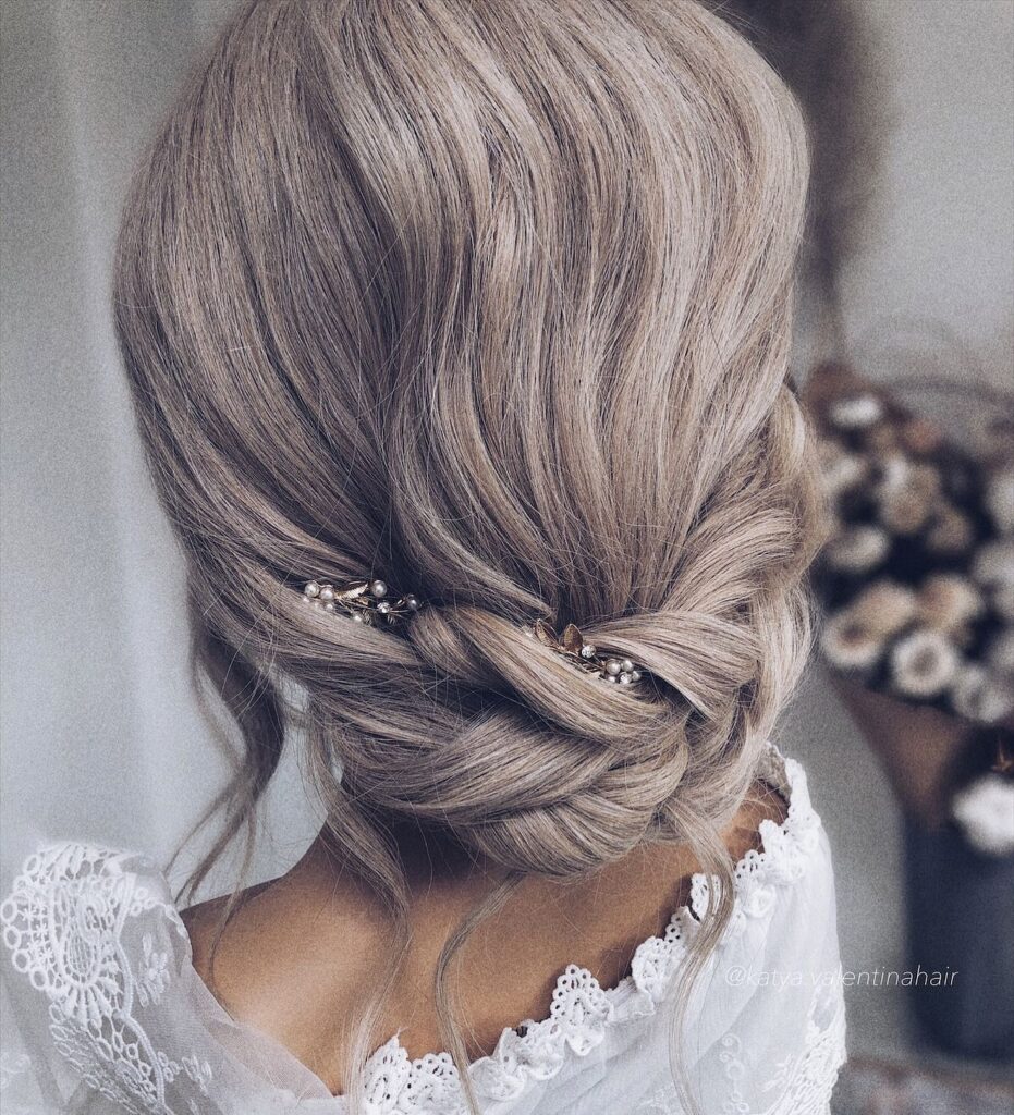 Soft blonde twisted low chignon with pearl hair accessory and loose face framing strands.