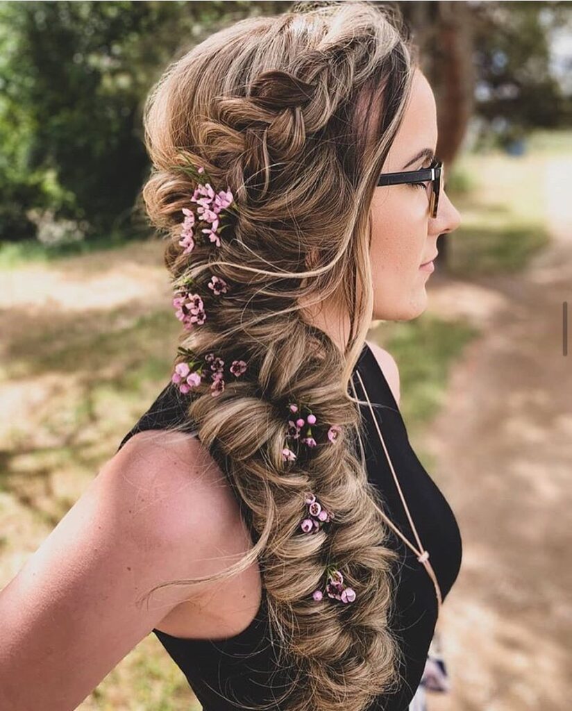 Loose side fishtail braid with soft waves and small pink flowers woven throughout.