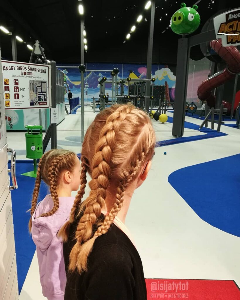 Two girls wearing chunky blonde braided hairstyles in an indoor activity space.