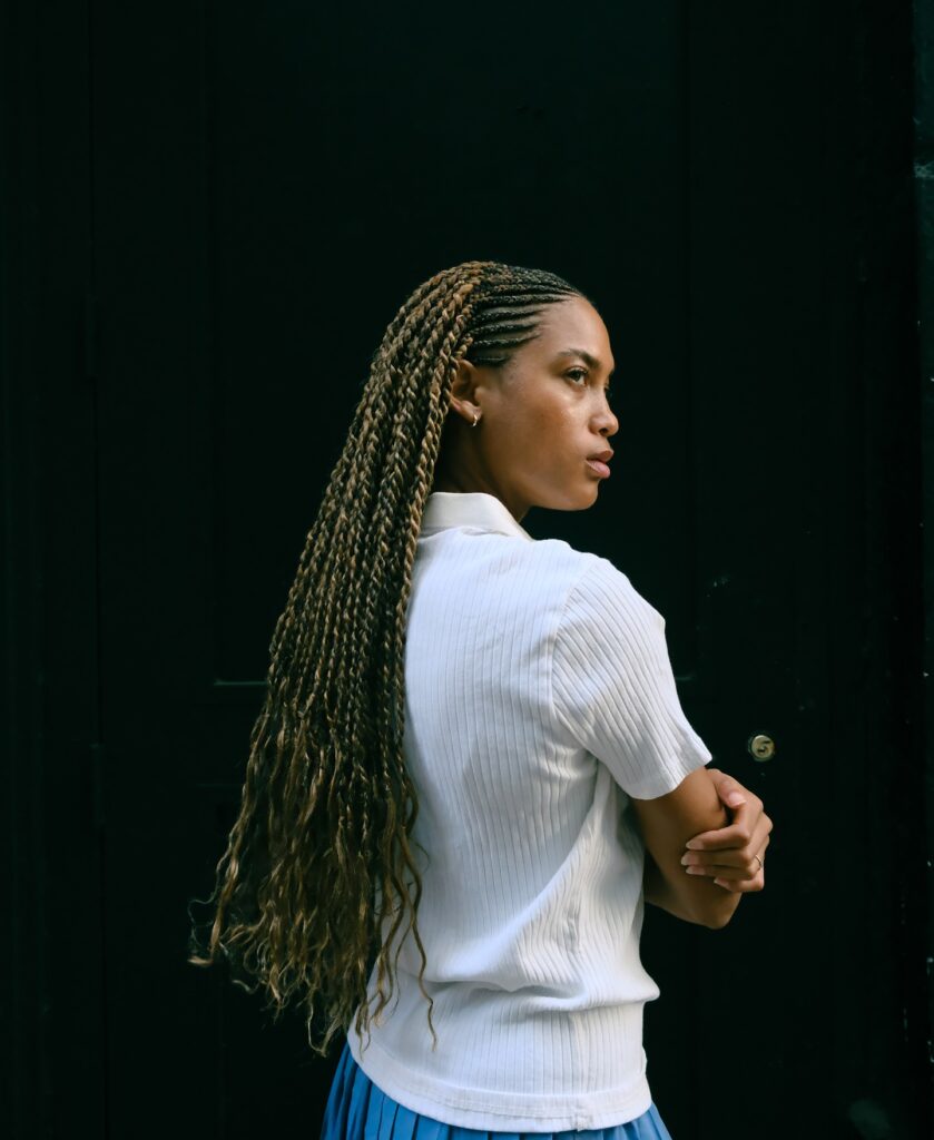 A woman with long feed in braids and soft wavy ends standing against a dark background.