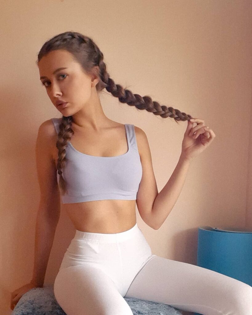 A woman with long brunette braided pigtails wearing light workout clothes and holding one braid to the side.
