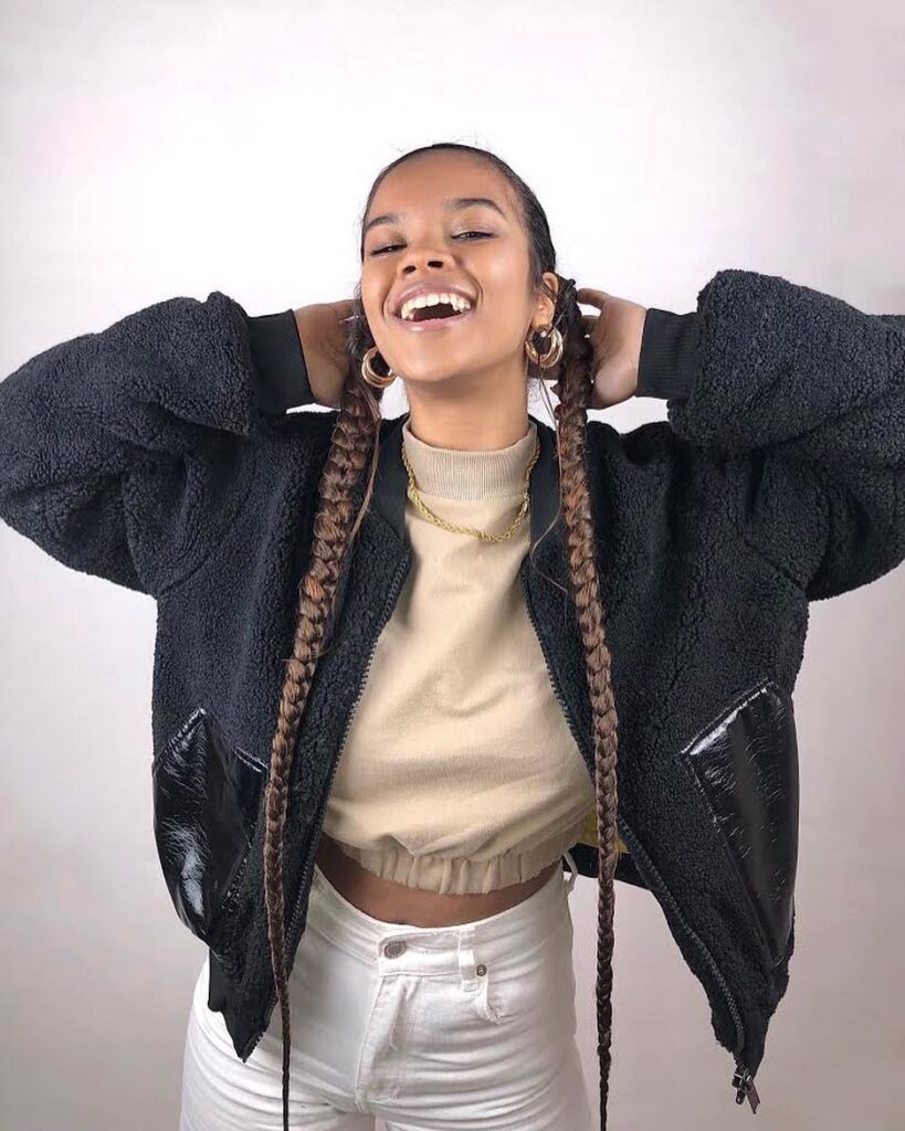 A woman smiling with two long brown braided ponytails and a middle part.