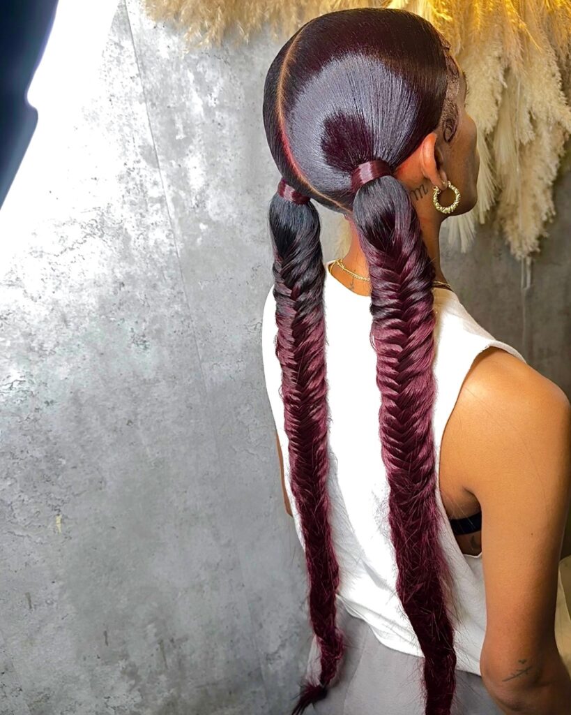 A woman with sleek burgundy fishtail braided ponytails and a middle part.