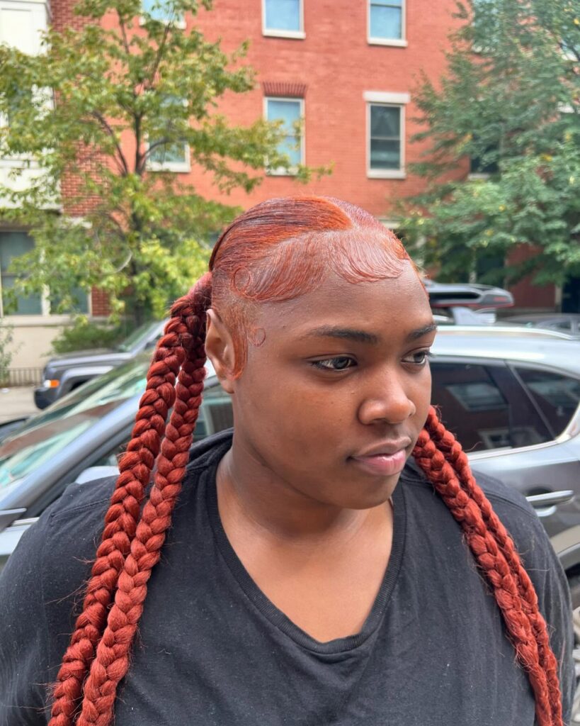 A woman with sleek copper braided ponytails and styled baby hairs.