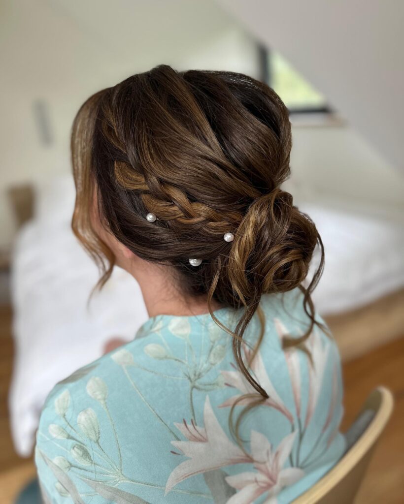 Loose brunette braided low bun hairstyle with pearl accents and soft curled strands.
