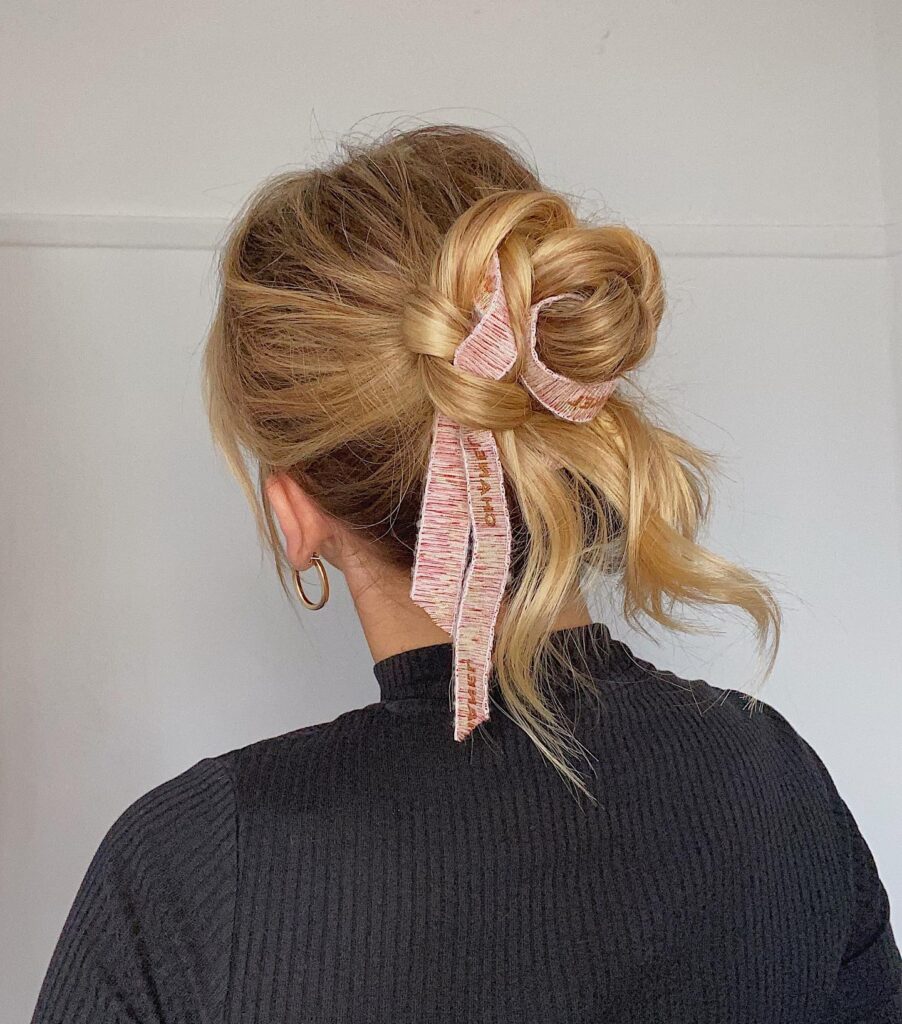 Messy blonde bun hairstyle with a ribbon accessory and soft wispy texture.