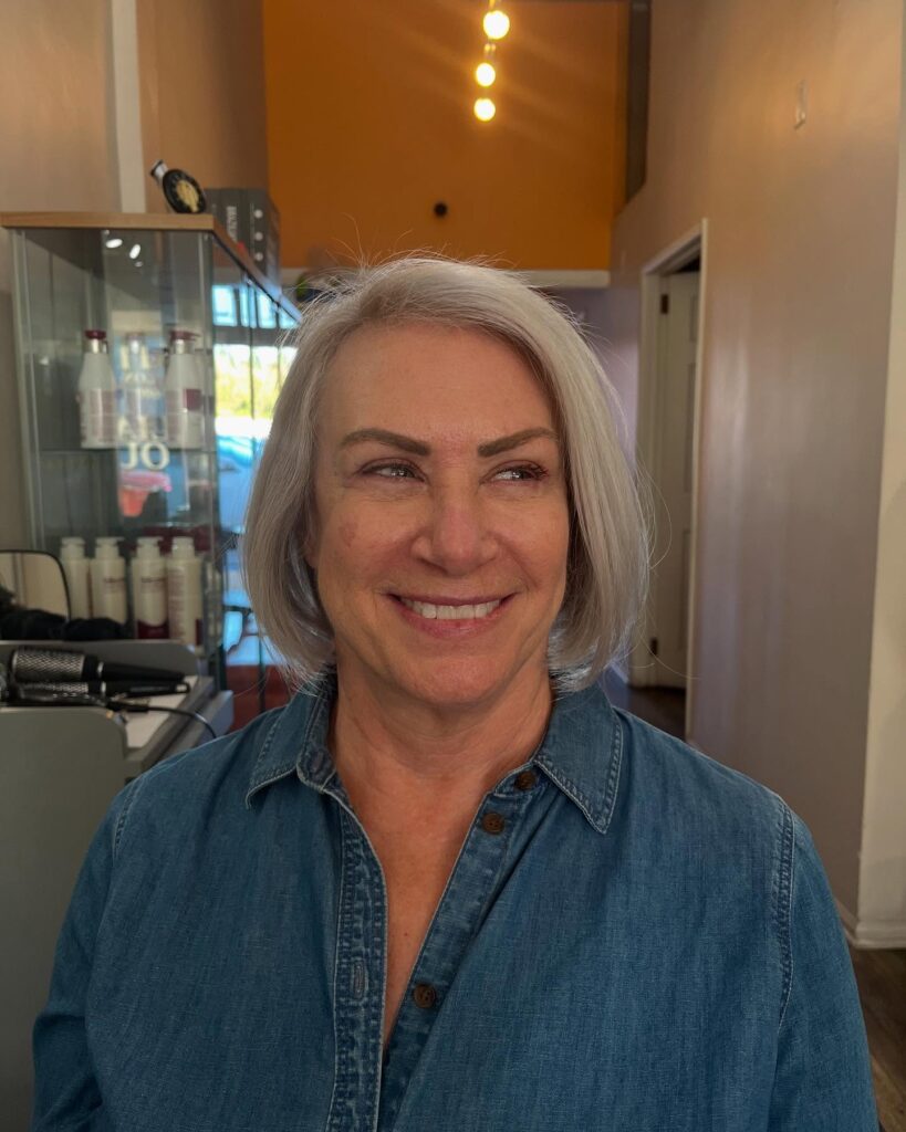 A woman with a square face wears a medium length ashy blonde hairstyle with voluminous layers and ends that curve toward her face.