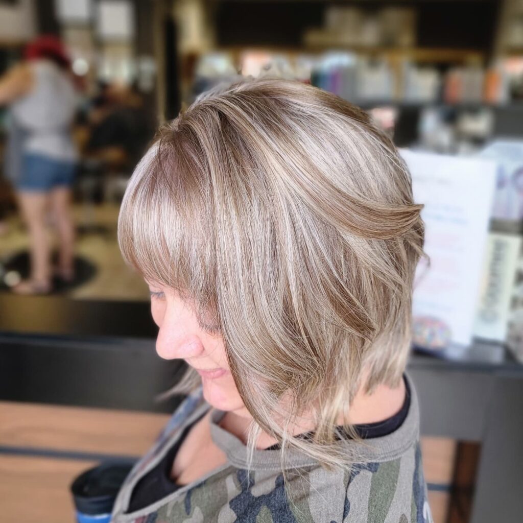 A woman with a square face shape wears a chin-length ash blonde bob with textured layers and full bangs.