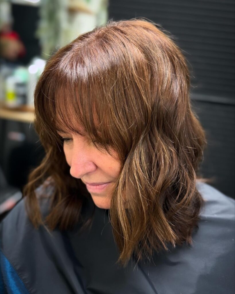 A woman with a square face shape showcases a shoulder-length chestnut brown hairstyle with messy waves and full wispy bangs.