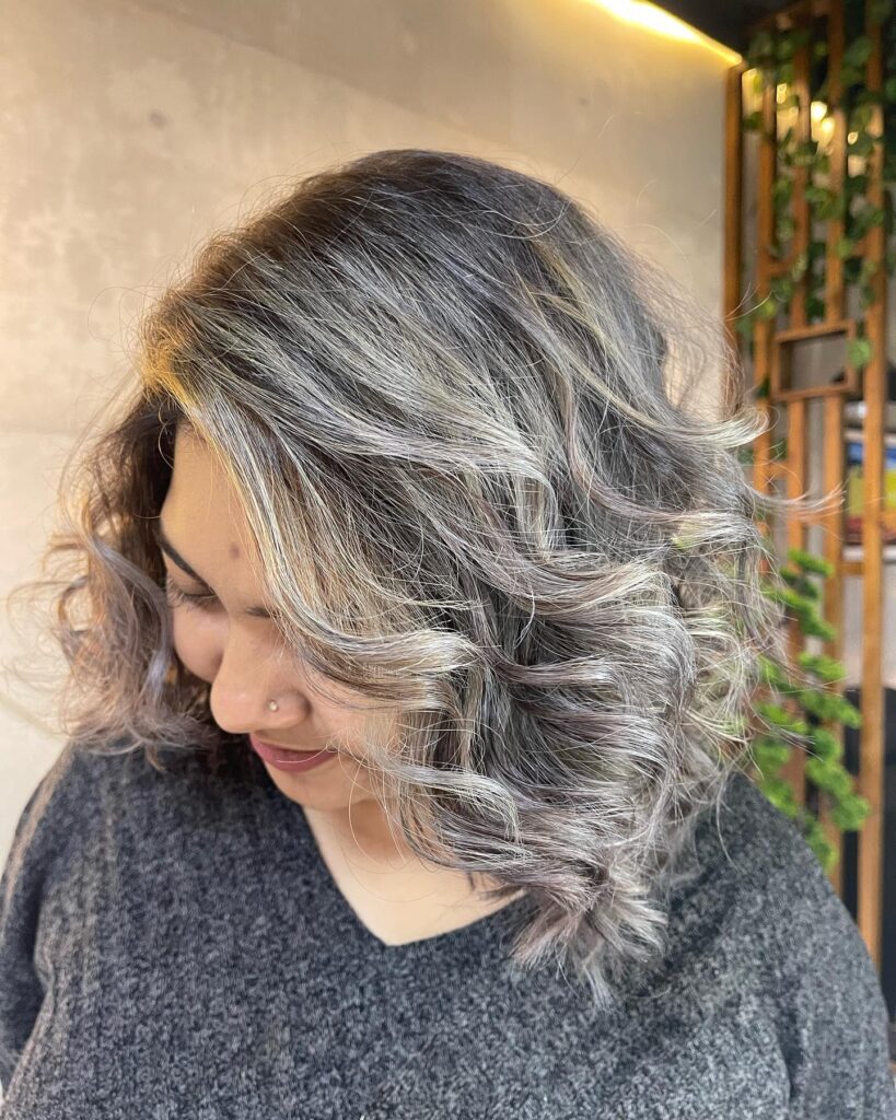 A woman with a square face shape wears a curly shoulder-length salt and pepper hairstyle with silver highlights and a side part.