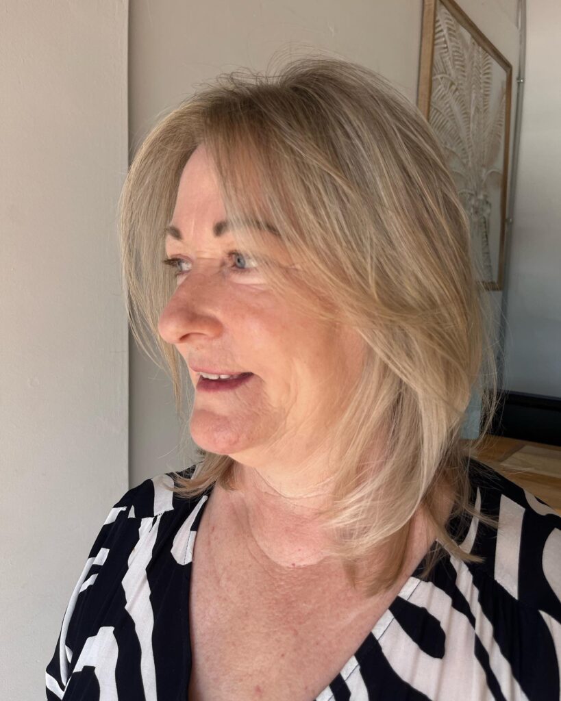 A woman with a shoulder length honey blonde hairstyle featuring soft layers and wispy face framing bangs.