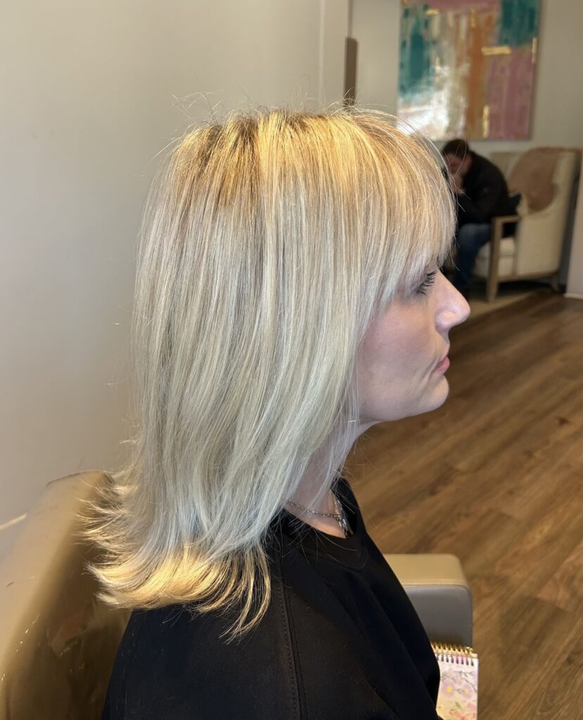 A woman with a mid length champagne blonde hairstyle featuring straight hair, a full fringe, and outward flicked ends.