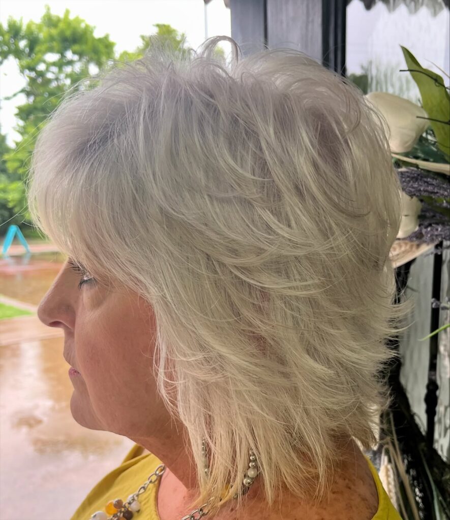 A woman with a round face displays a short tapered silver hairstyle with textured height on top and soft feathery layers.