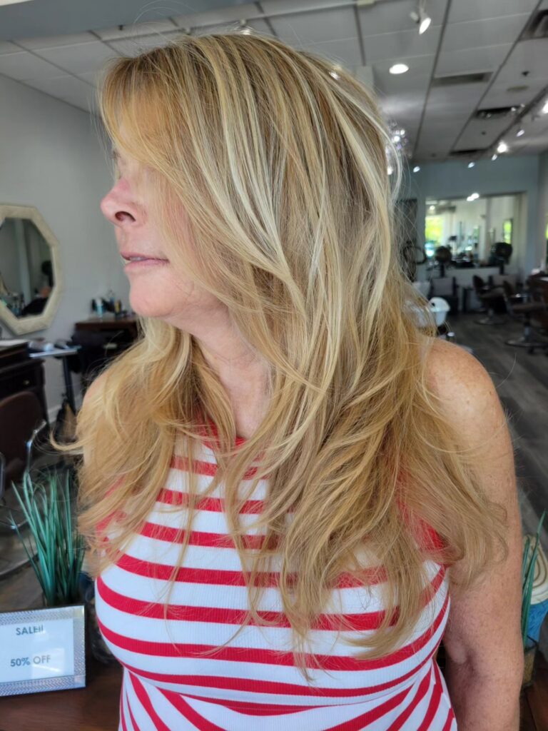 A woman with long golden blonde hair styled in lots of layers and loose waves poses in a hair salon.