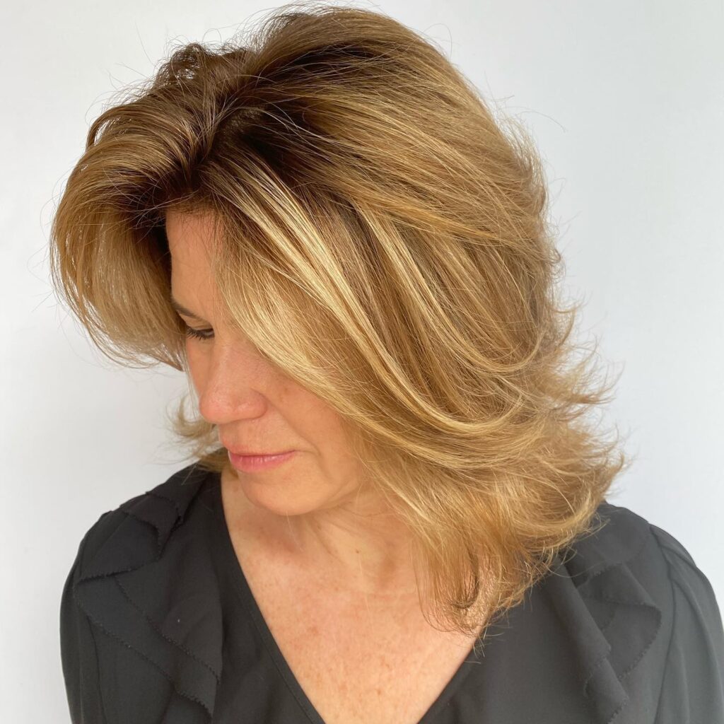 A woman with a round face wears a voluminous honey blonde lob with feathered layers and outward flicked ends.