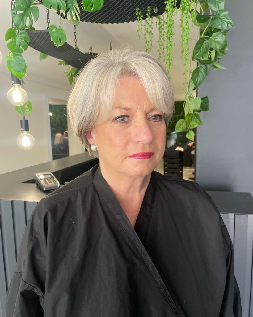 A woman with a round face shape wears a polished silver tapered bob with long side-swept bangs and soft layers.