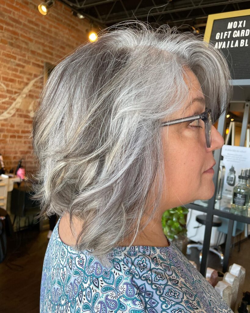 A woman with a round face shape wears a wavy salt and pepper lob with layered side swept bangs.