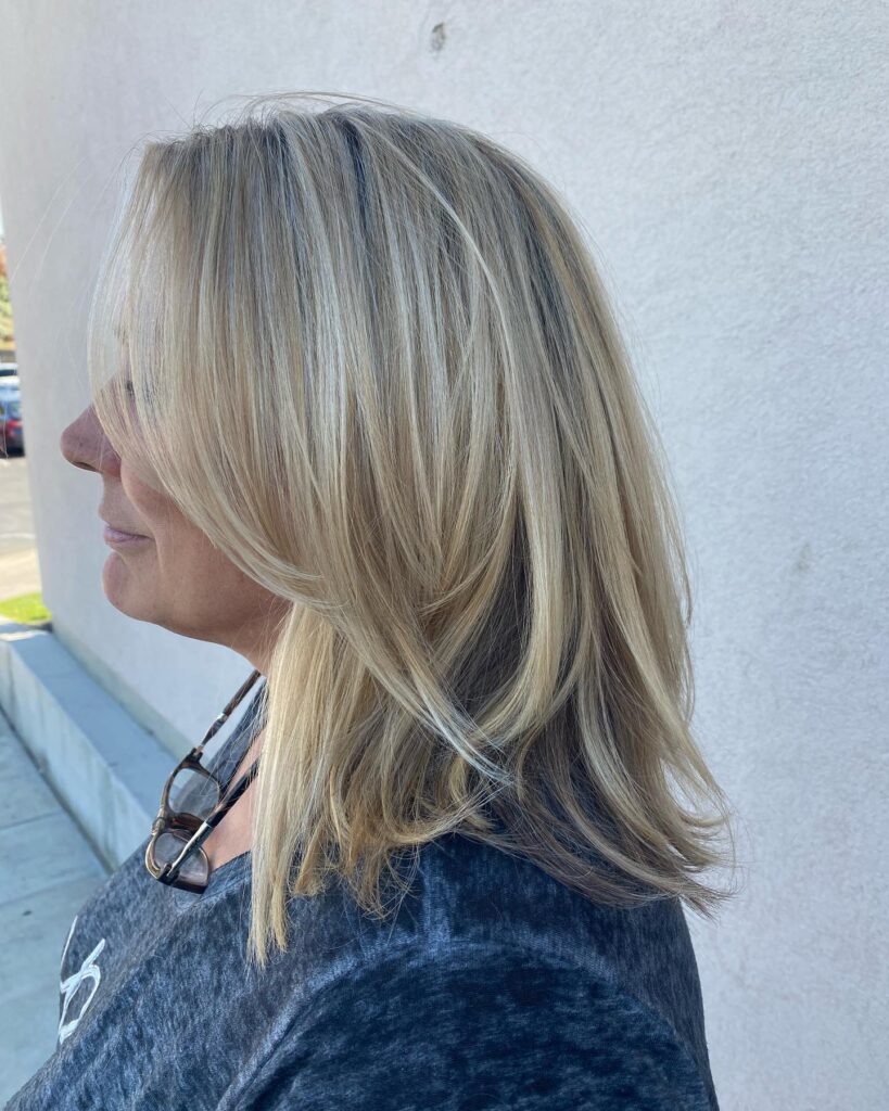 A woman with a round face displays a shoulder length honey blonde lob with feathered layers and bright highlights.