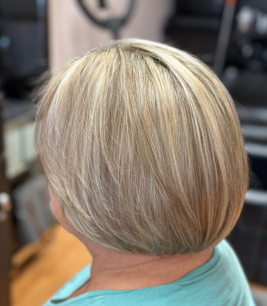 A back view of a woman with a rounded chin length bob featuring honey blonde highlights and a smooth straight texture.