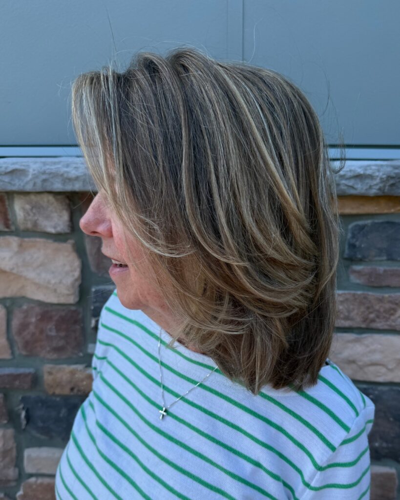 A woman with a round face displays a shoulder length ash brown lob with face framing layers and sandy blonde highlights.