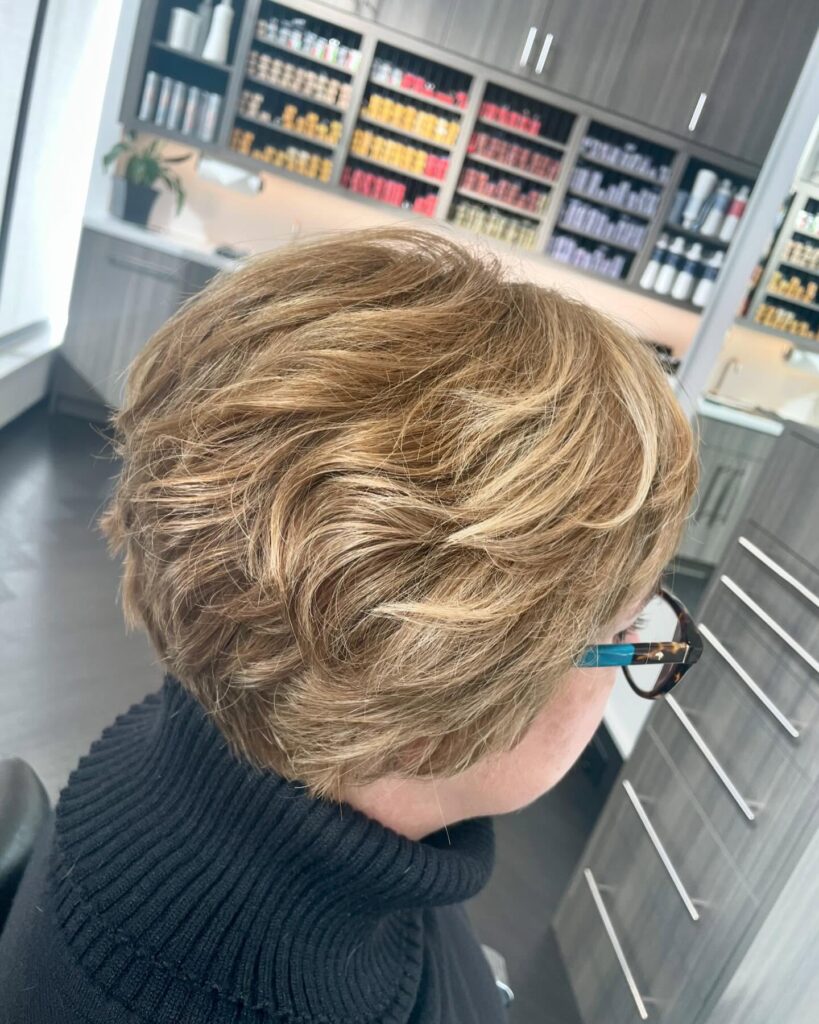 A woman with a short stacked pixie cut in honey blonde featuring soft wavy layers and volume at the back.