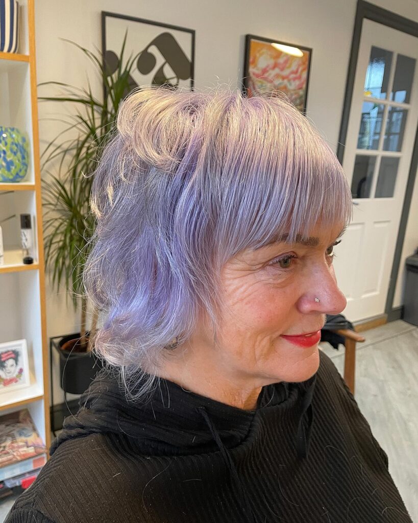 A woman with medium length lavender hair featuring a shaggy layered cut and a straight across fringe.