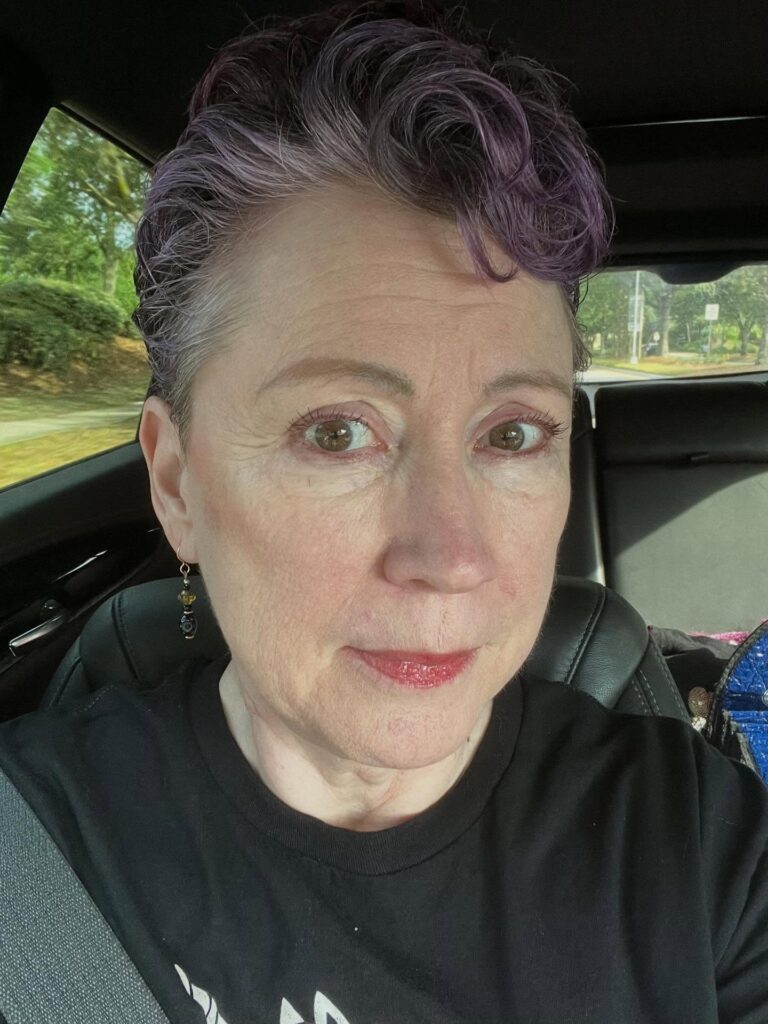 A woman with a short tapered pixie cut featuring natural grey roots and dusty violet waves on top.