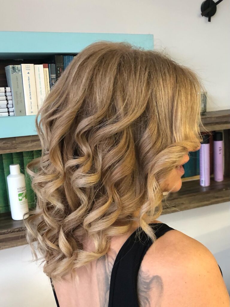A woman with medium length sandy blonde hair styled into tight bouncy ringlet waves.