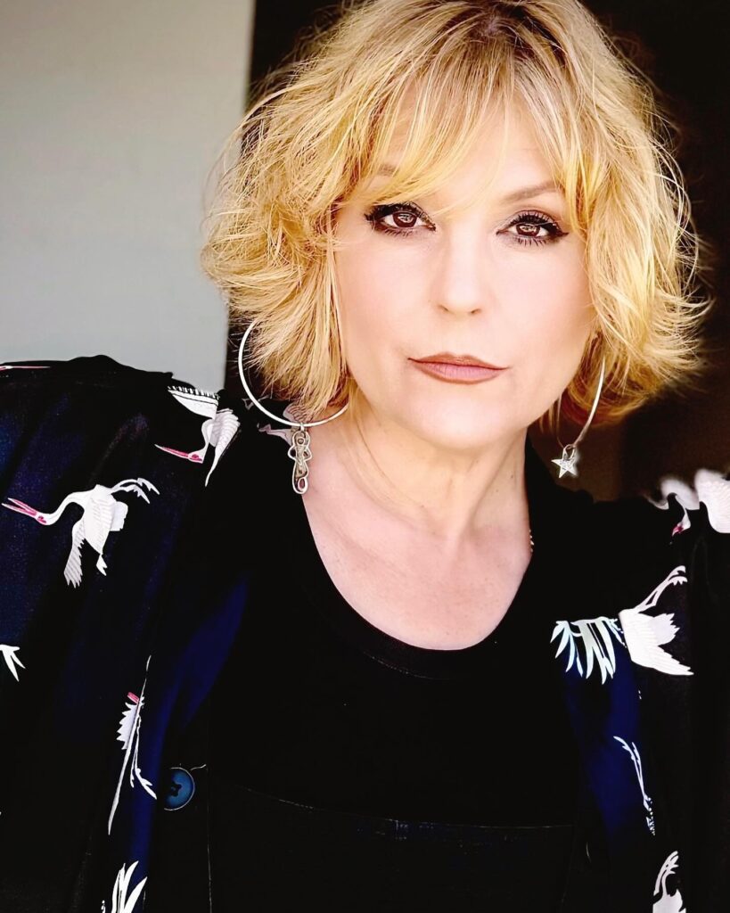 A woman with a chin-length honey blonde bob styled in soft, tousled waves with a wispy side-swept fringe.