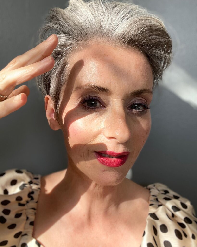 A woman with a sleek silver pixie haircut and red lipstick poses in sunlight.