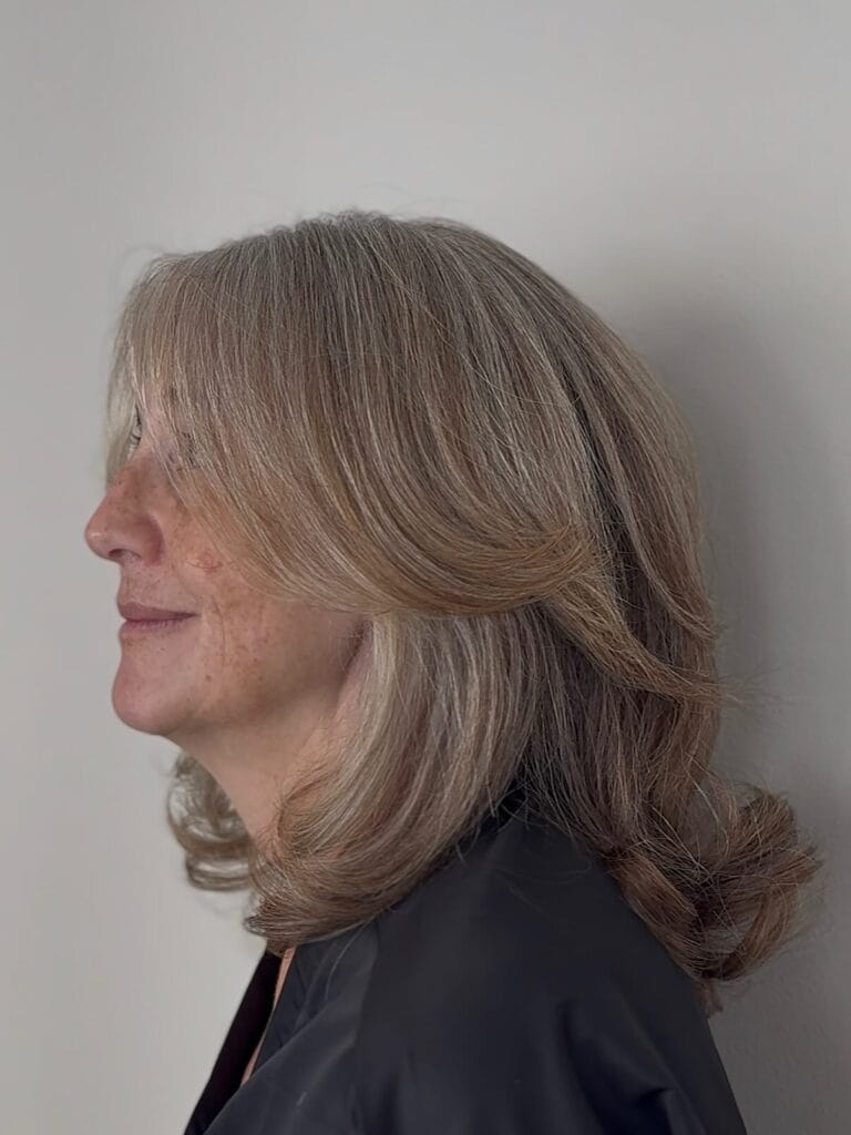 A woman with ash blonde and silver hair displays a shoulder length layered haircut with face framing pieces and flicked out ends.