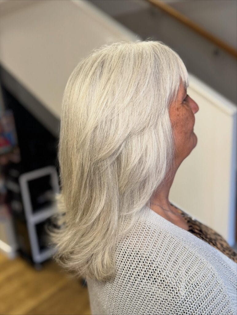 A woman with bright silver hair displays a long layered haircut featuring a soft wispy fringe and shoulder length ends.