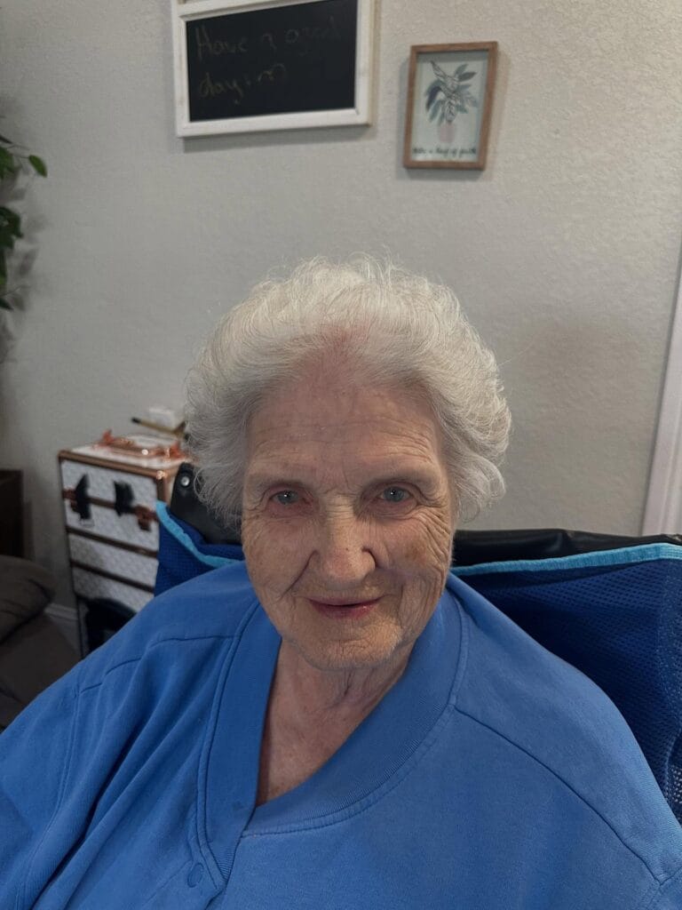 An elderly woman with voluminous bright white hair is smiling while wearing a blue shirt.