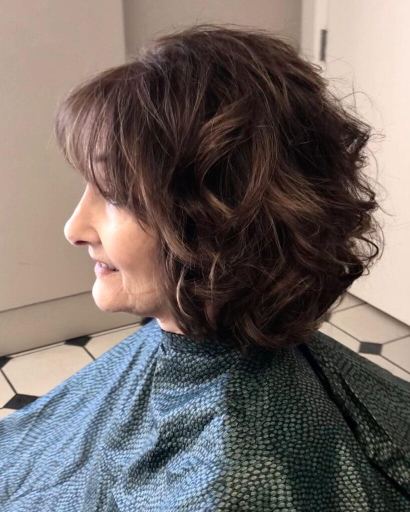 A woman from a side profile wearing a voluminous curly chocolate brown bob with light wispy bangs.
