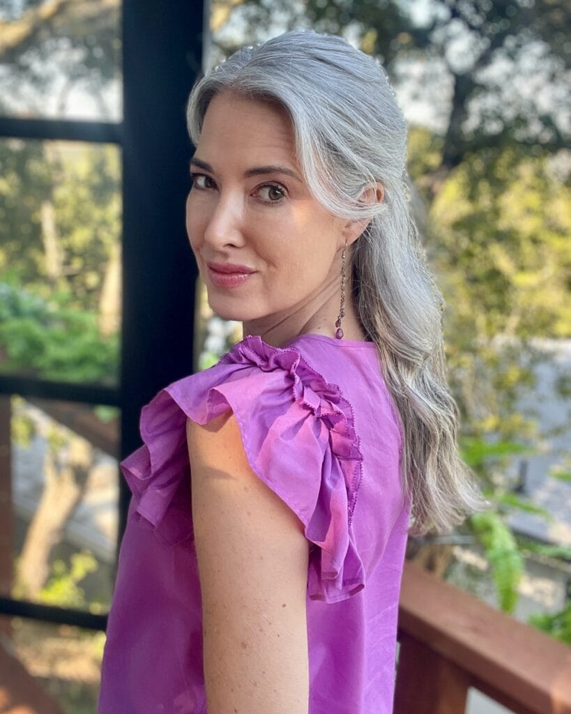 A woman with long wavy silver hair wears a half up half down style while looking over her shoulder.