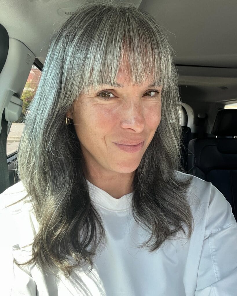 A woman with long silver and grey hair featuring a full straight across fringe and soft natural waves.