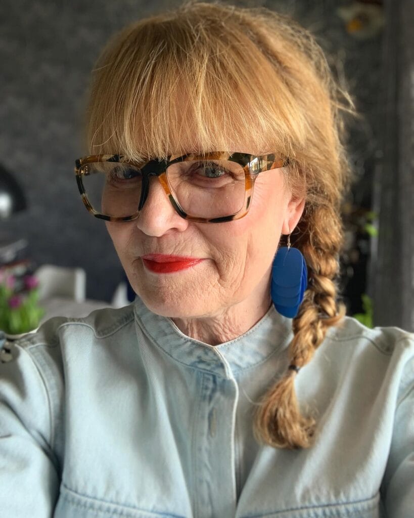 Long blonde hair in a side braid with straight full bangs.